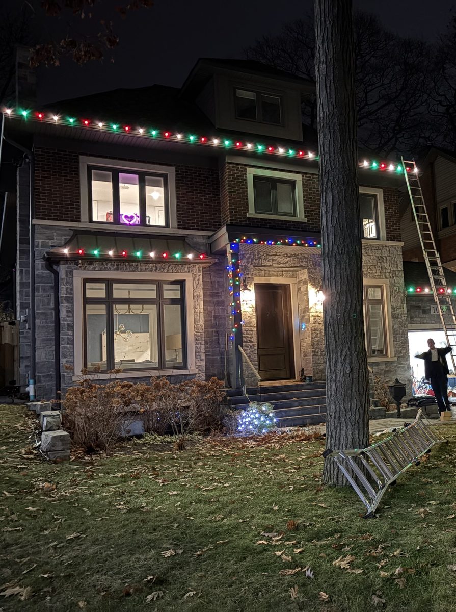 The same house with red, green and white lights on the roofline and porch, crew still on the ladder