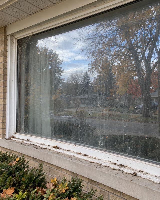 Large picture window on a beige brick bungalow, glass dusty and streaked before cleaning